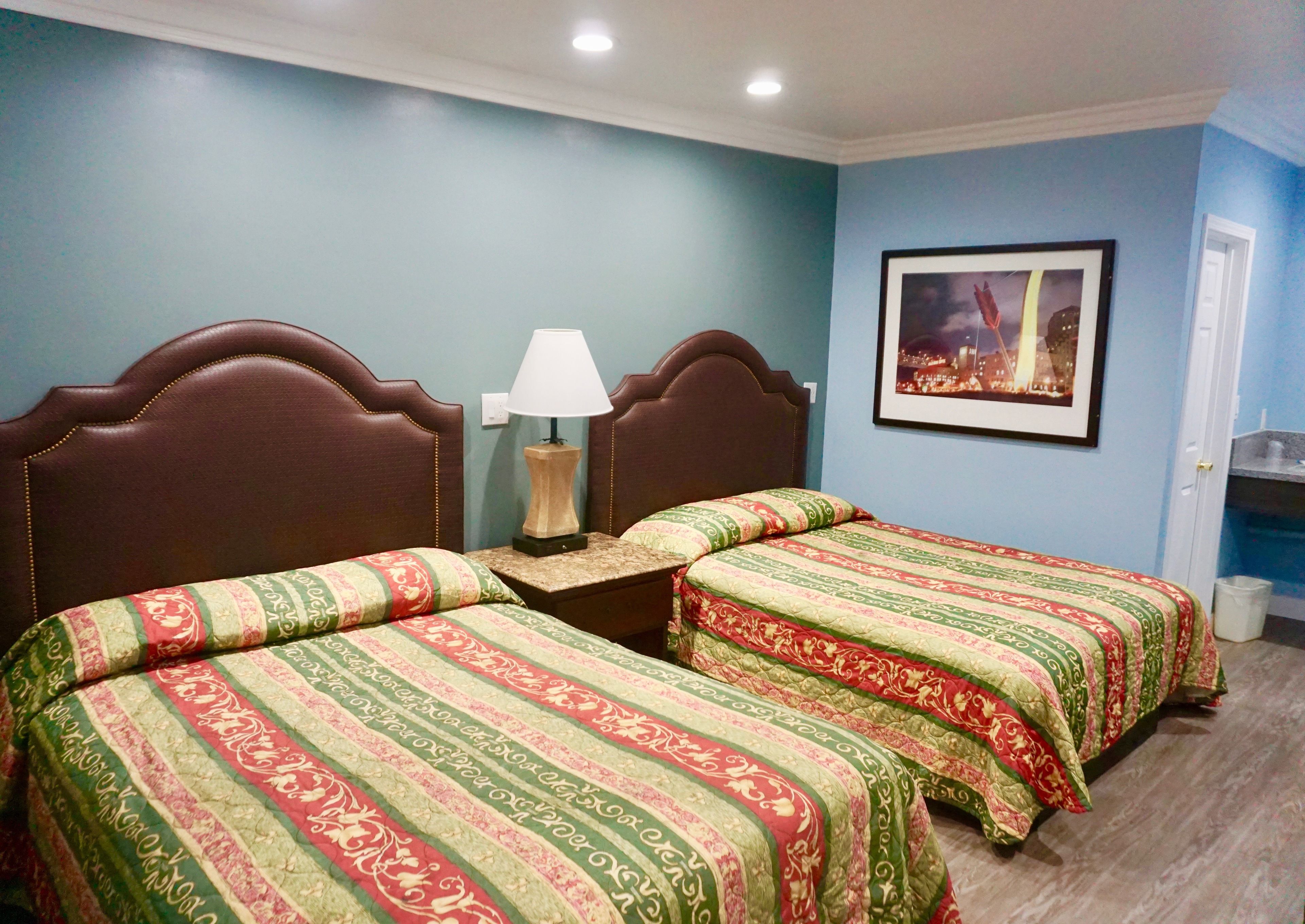 A room with two beds, a nightstand with a lamp, a framed picture on the wall, and a door to the right.