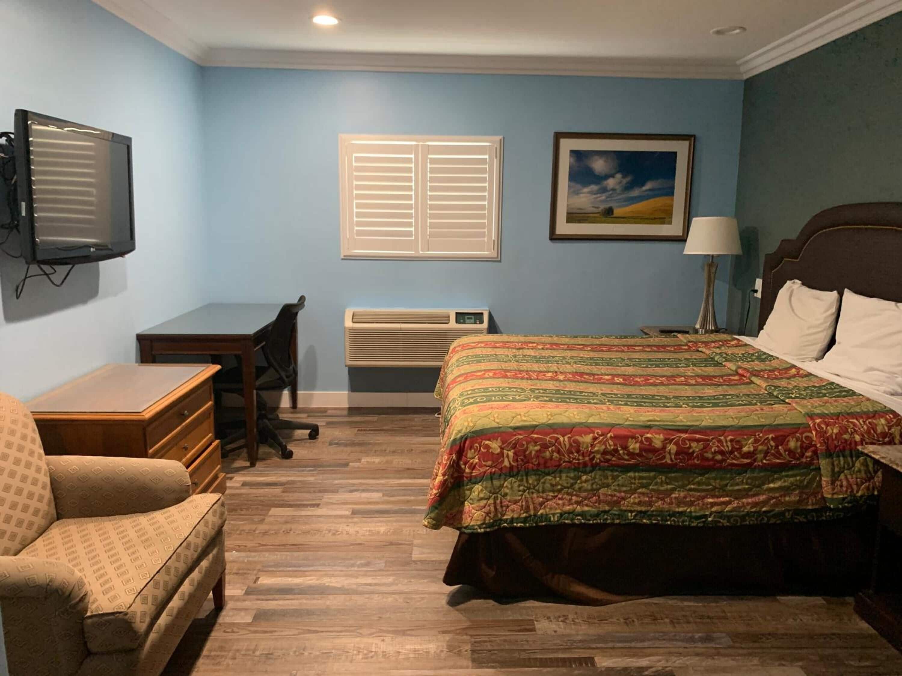 A bedroom with a bed, a desk, a chair, a television, and a window with blinds.