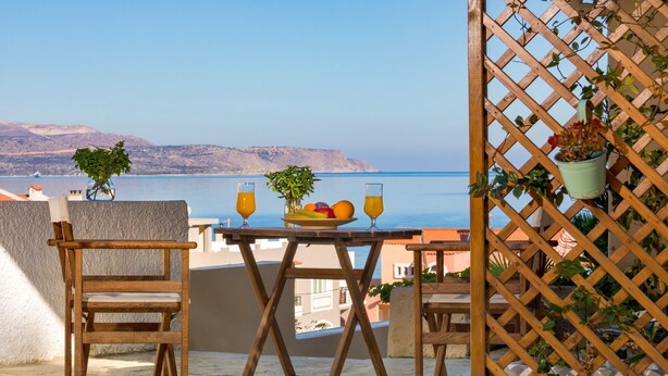 Et træbord med to stole, en tallerken med frugt og to glas appelsinjuice på en balkon med udsigt over havet.