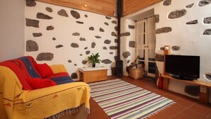 A cozy living room with a stone wall, a sofa with a yellow blanket, a TV on a wooden stand, and a fireplace. - A small cottage in the center of Horta, Faial Island, Azores (Horta)