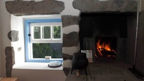 A stone fireplace with a visible fire, a window with a blue frame, and a stone wall. - A small cottage in the center of Horta, Faial Island, Azores (Horta)