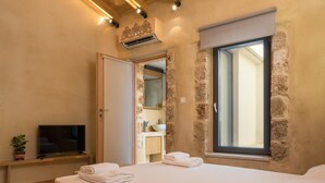 A bedroom with a wooden bed, a TV on a shelf, a bathroom with a stone wall, and a window with a view. - Seaside 2 floor Maisonette with Jacuzzi (Χανιά)