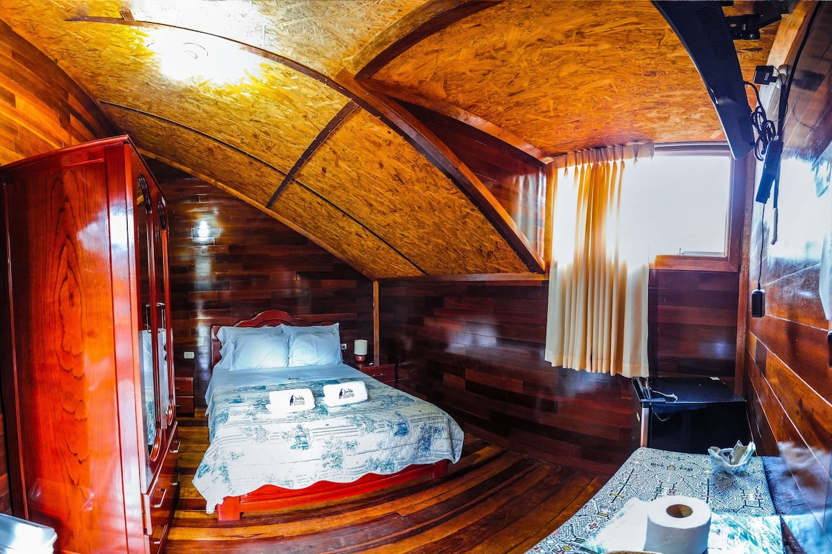 A wooden cabin room with a bed, a small table with a basket, and a window with curtains.