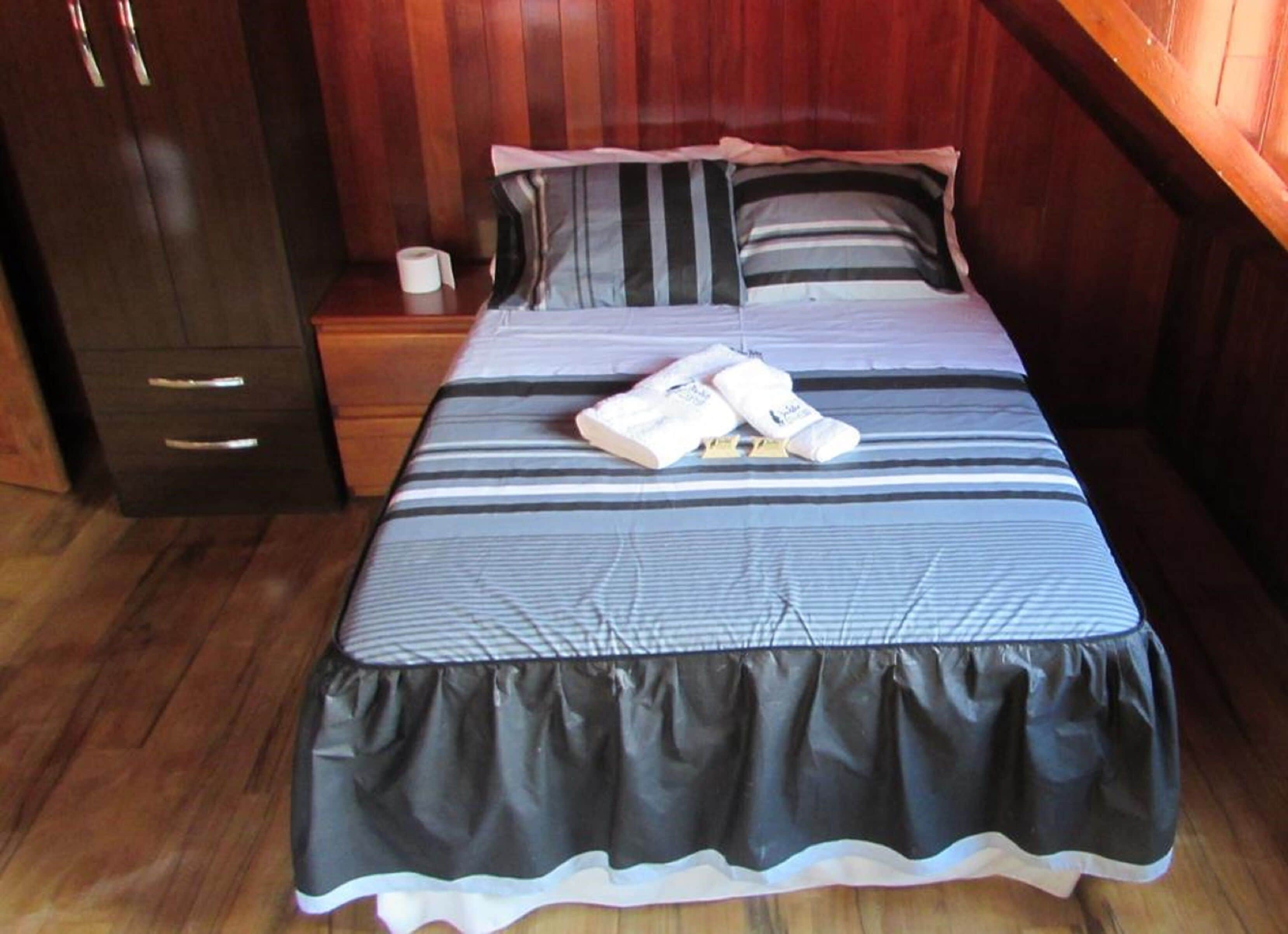 A neatly made bed with striped bedding, a wooden nightstand with a roll of toilet paper, and a dark wooden wardrobe.