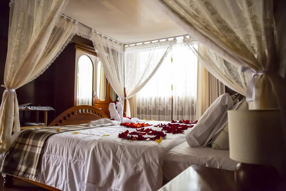 A four-poster canopy bed with white linens and rose petals, a wooden headboard, and a bedside table with a lamp.