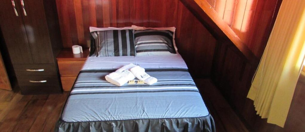 A neatly made bed with striped bedding and a dark-colored skirt, a wooden nightstand with a cup, and a wooden wardrobe.