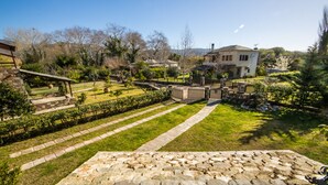 A residential area with a stone pathway leading to a house with a white facade and a chimney. There are trees and a garden with a wooden fence.