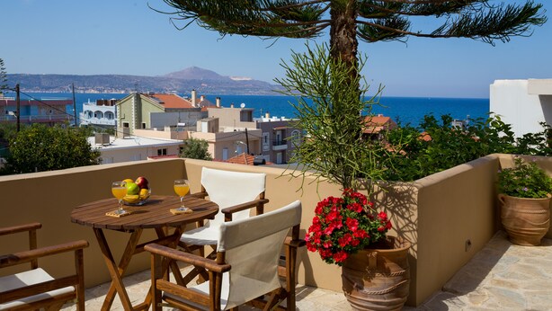 A terrace with a wooden table and chairs, a potted plant, and a view of the sea and buildings. - Avra Apartments, Maistros (Καλύβες)