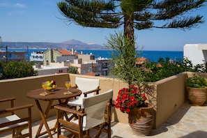 Une terrasse avec une table et des chaises en bois, un pot de fleurs et une vue sur la mer et les bâtiments.
