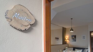 A cozy living room with a wooden sign reading "Maistros," a dining table with chairs, a gray sofa, and a hanging light fixture. - Avra Apartments, Maistros (Καλύβες)
