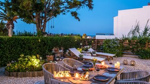 An outdoor dining area with a table set for two, surrounded by wicker chairs, and a fire feature in the center.