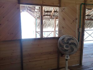 A room with wooden walls, a fan, and a window with a thatched roof visible through the window. - Dahlandia (Isla Mucura)