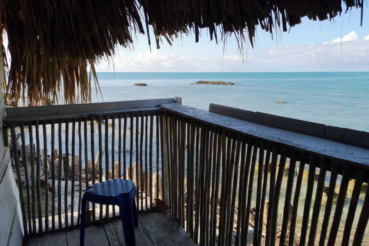 Eine Holzterrasse mit blauem Stuhl, mit Blick auf einen Strand und ruhiges Wasser sowie in der Ferne Felsen.