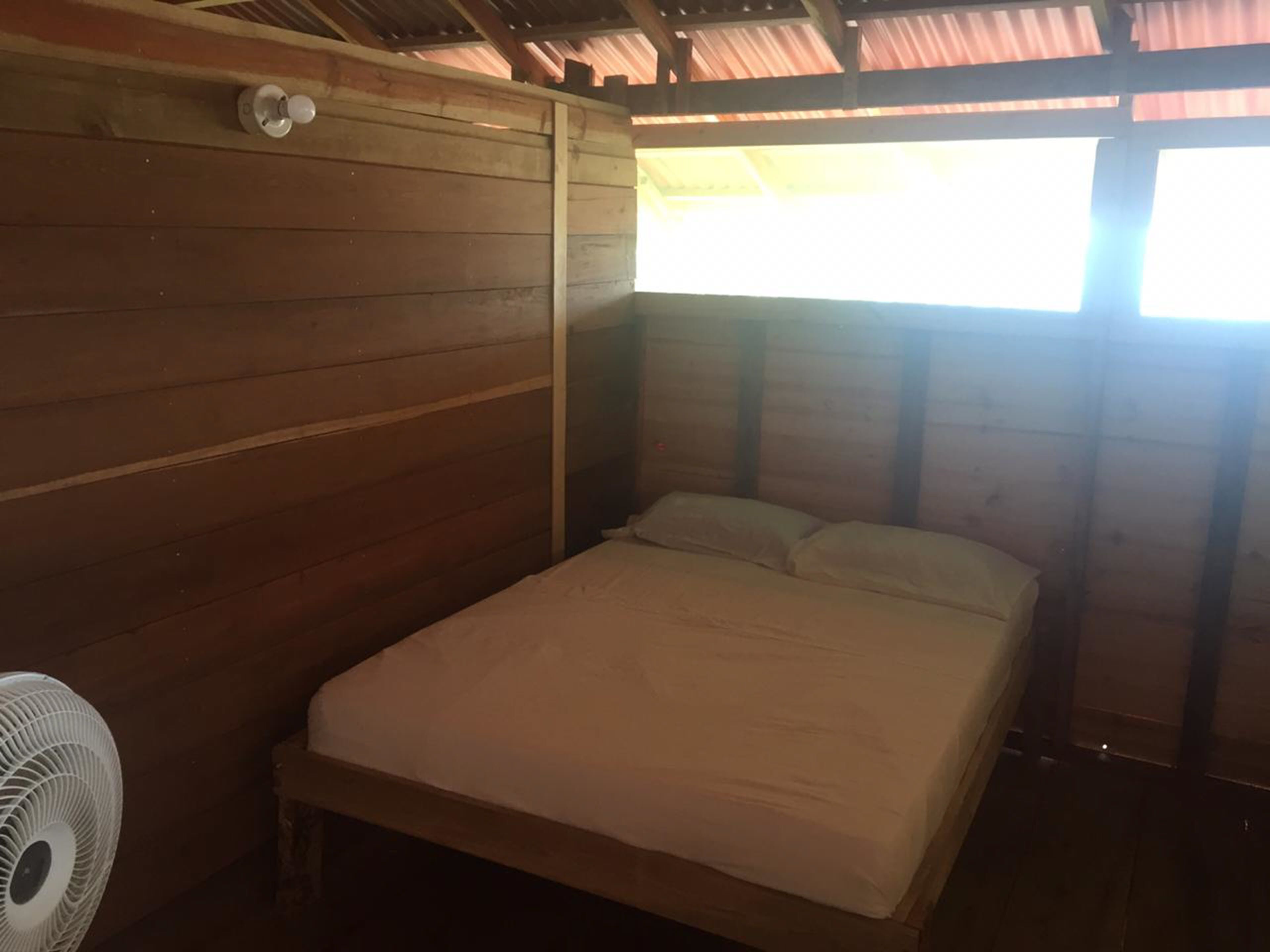 Una habitación pequeña con cama individual, paredes de madera y techo inclinado. Un ventilador blanco a la izquierda y una ventana a la derecha.