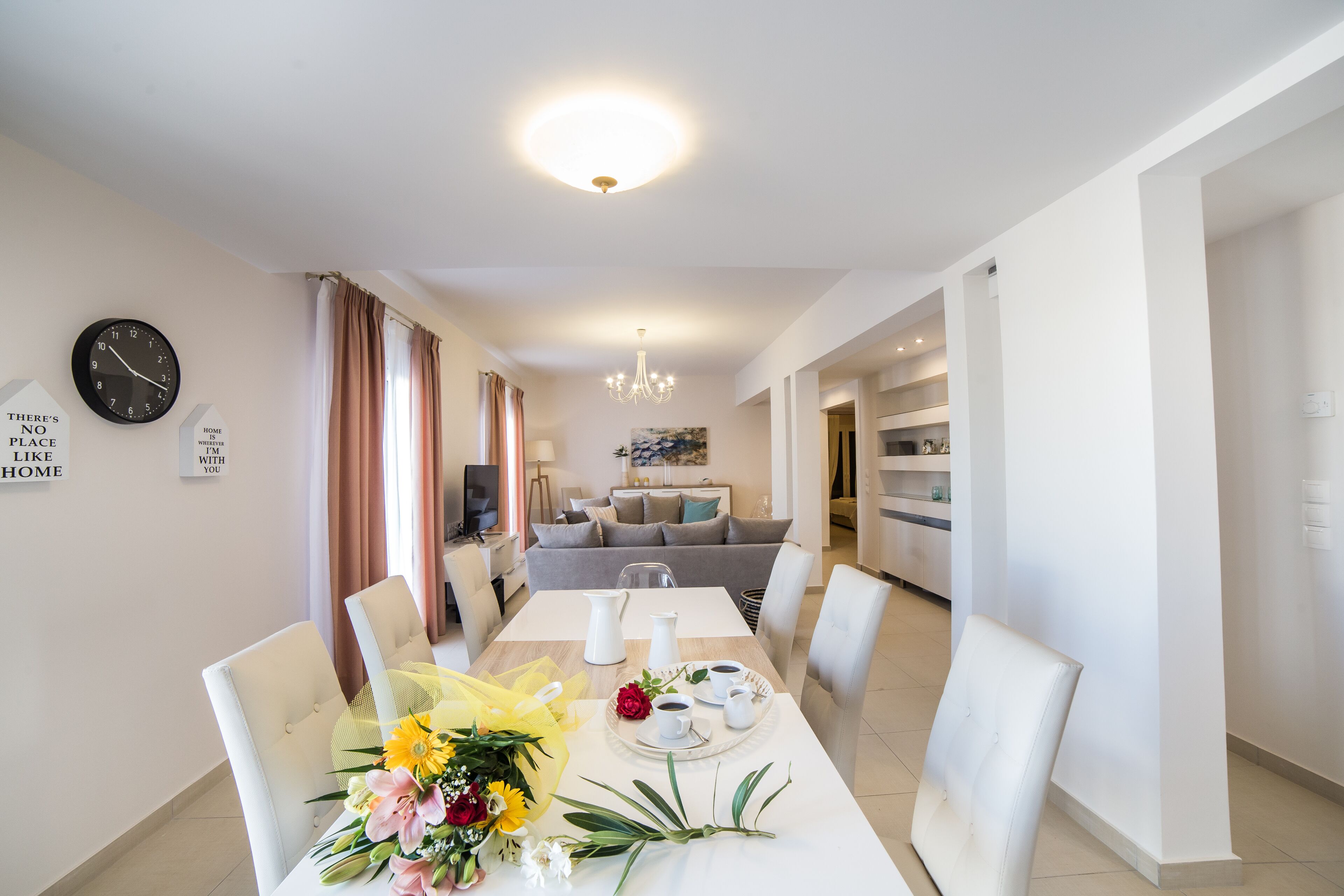 Un espace repas moderne avec une table blanche dressée pour deux personnes, une horloge au mur et une vue sur le salon.