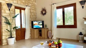 A cozy living room with a television, a dining table with a book and wine glass, a fruit basket, and a potted plant. - Eros villa ,private Pool,short drive to beach (Rethymno)