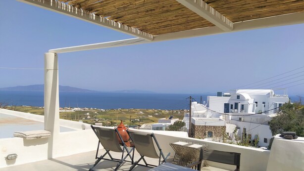 Terraço com mesa e cadeiras, com vista para uma cidade costeira de edifícios brancos e o mar.