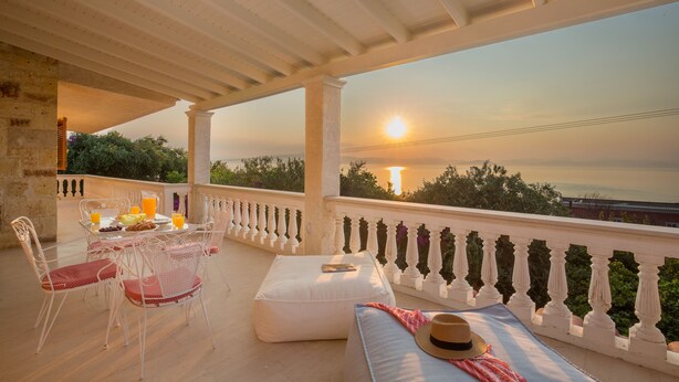 Une terrasse extérieure avec une table dressée pour le petit-déjeuner, des sièges blancs confortables et une vue sur le coucher de soleil sur la mer.