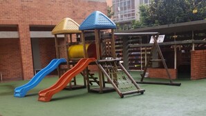 A playground with slides and climbing structures, surrounded by a brick building and a green artificial turf area.