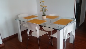 A glass-top dining table with four white chairs, a potted plant with yellow flowers, and a white wall.