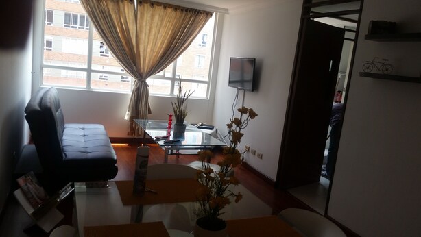 A living room with a sofa, a glass coffee table, a TV mounted on the wall, and a window with curtains.