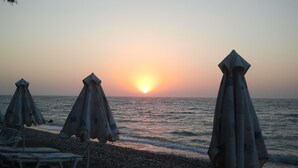 A beach with lounge chairs and umbrellas, a sunset in the background, and a rocky shoreline. - Two bedroom apartment only 500 meters to the beach (Ιαλυσός)