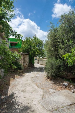 A narrow concrete pathway leading to a stone gate, surrounded by dense greenery and a clear blue sky.