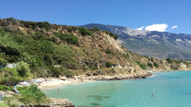 Ein Küstenstrand mit klarem türkisblauem Wasser, Felsformationen und einem bergigen Hintergrund.