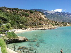 A coastal beach with clear turquoise water, rocky outcrops, and a mountainous backdrop.