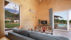 Ein gemütliches Wohnzimmer mit einer großen Couch, einem Flachbildfernseher und einem Couchtisch. Ein Fenster bietet Blick auf einen Pool und Berge.