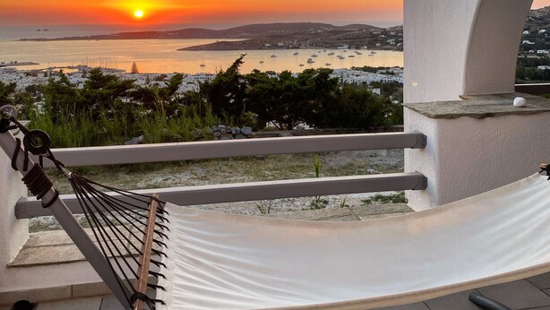 Een hangmat op een balkon met uitzicht op een zonsondergang, bootjes en een stad in de verte.