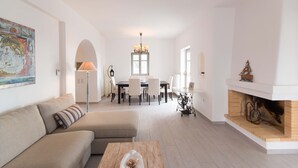 Een ruime woonkamer met een bank, salontafel en open haard. Op de achtergrond is een eethoek met een tafel en stoelen te zien.