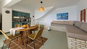 Una cocina moderna con armarios azul verdoso, una zona de comedor con una mesa de madera y sillas de mimbre, y una sala de estar con sofá y vista al mar.
