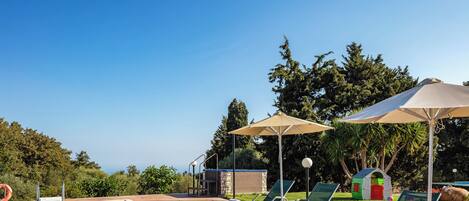 A swimming pool with lounge chairs and umbrellas, surrounded by greenery and a play area.
