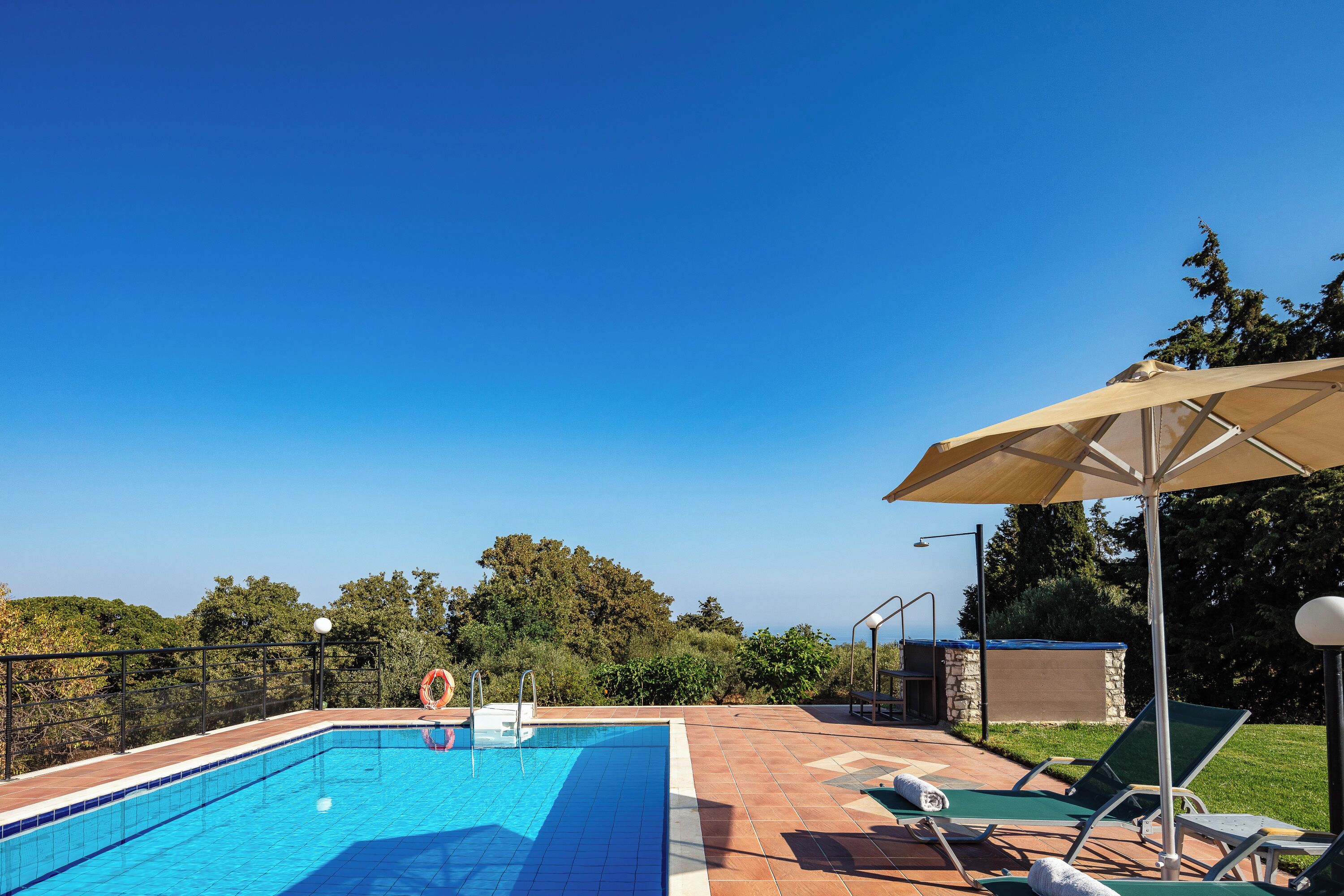 A rectangular swimming pool with clear blue water, surrounded by a tiled deck. There are lounge chairs and a large umbrella providing shade.