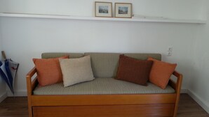 Un sofá de madera con cojines a rayas y dos almohadas, una pared blanca con un estante arriba, y un suelo de madera.