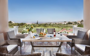 Une terrasse meublée de mobilier en osier, une table dressée pour deux personnes, un panier de fruits et une bouteille de vin.