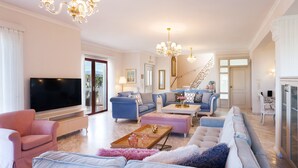 A spacious living room with a large flat-screen TV, a blue sofa set, a pink armchair, and a staircase leading to another level. - Villa Vanelia Mare - Guest Favorite Elegant Stay! Authentic Greek Hospitality! (Χανιά)