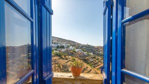 A view through blue-framed windows overlooking a hillside village with white buildings and a potted plant on the ledge. - "Art House"          (Τήνος)