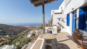 A traditional white building with blue doors and windows, a stone table, and a view of the countryside. - "Art House"          (Τήνος)