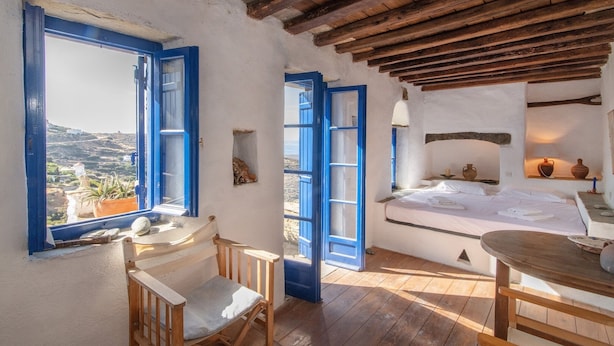 A room with a wooden ceiling, a bed with white linens, a table with a chair, and a view of the landscape through a window with blue shutters. - "Art House"          (Τήνος)