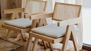 Dos sillas de madera con respaldos trenzados y asientos acolchados, ubicadas sobre un piso de baldosas con una cortina al fondo.
