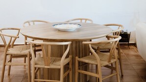 Un comedor con una mesa redonda de madera, seis sillas de mimbre y un cuadro enmarcado con un paisaje costero en la pared.