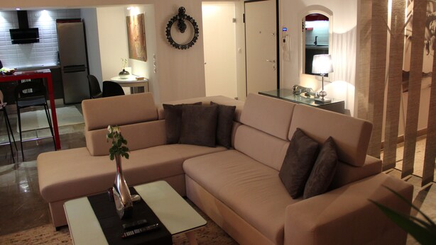 Una sala de estar con un sofá seccional color beige, una mesita con un florero, y una zona de cocina al fondo.