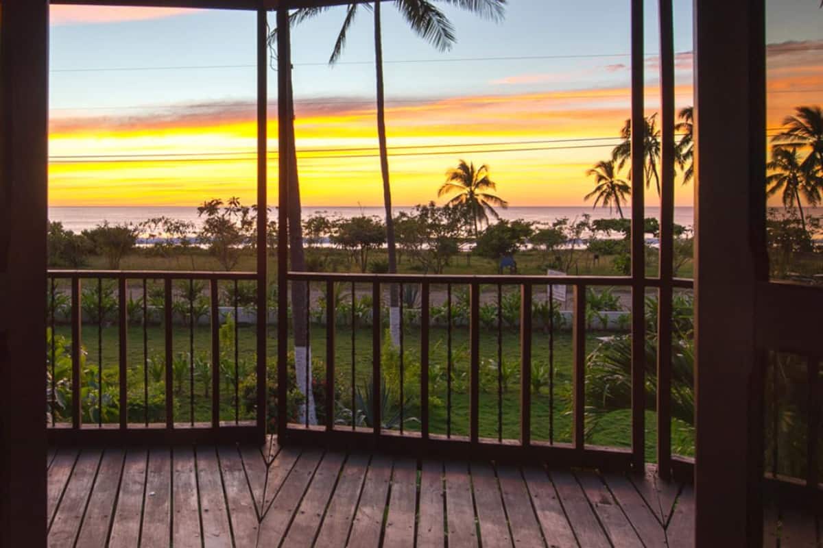 A wooden deck with a view of a sunset over the ocean, palm trees, and a grassy area.