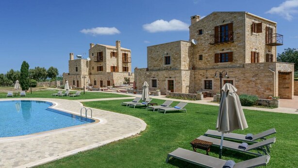 Un espace aménagé au bord de la piscine, avec des chaises longues et des parasols, des bâtiments en pierre dotés de balcons, et un ciel d’un bleu limpide.