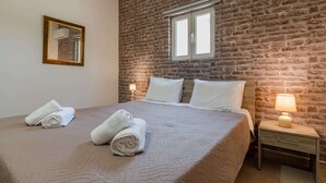 Habitación de hotel con pared de ladrillo, cama grande con sábanas blancas, mesita de noche con lámpara y ventana con persianas.