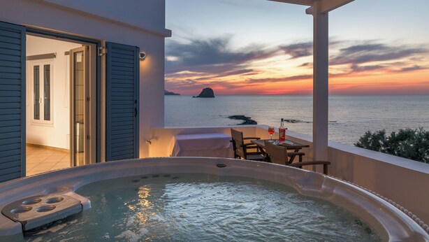 Uma varanda com banheira de hidromassagem, mesa e cadeiras, com vista para um pôr do sol sobre o mar.