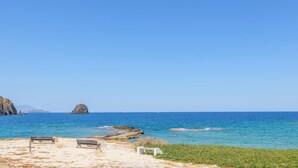 Uma praia com duas espreguiçadeiras, um afloramento rochoso e águas azuis calmas.