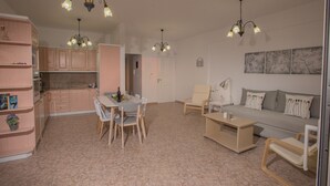 Een moderne woonkamer met een bank, salontafel en een eethoek met stoelen. Er is een keuken met kasten en een kroonluchter.